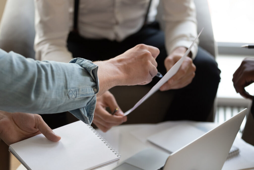 Close-up of a group of people sitting around a table engaged in discussion, with one person holding a document and pointing at it while another holds a notepad. A laptop and more papers are visible on the table, suggesting a collaborative meeting or brainstorming session.