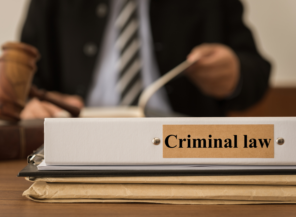 A close-up of a binder labeled "Criminal law" on a desk, with legal documents underneath. In the background, a person in a suit and tie is seen holding a book or paper, with a wooden gavel visible on the side, suggesting a courtroom or legal setting.